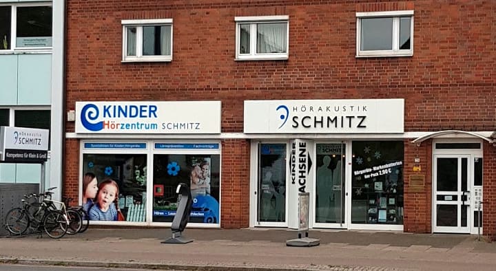 Fassade eines Hörakustik- und Kinderhörzentrums mit Schaufenstern, Beschilderung „Kinder Hörzentrum Schmitz“ und „Hörakustik Schmitz“.