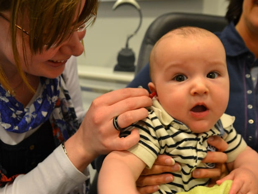 Eine lächelnde Frau setzt einem überraschten Baby ein kleines rotes Hörgerät ins Ohr, während eine weitere Person das Baby hält.